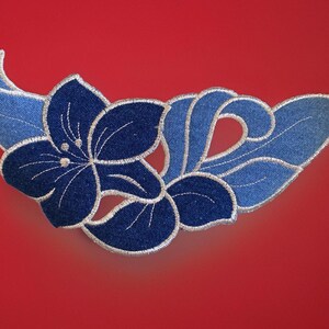 May include: A denim-style floral applique with a dark blue flower and light blue leaves and accents. The applique is embroidered with white thread and set against a red background. This decorative patch is suitable for clothing or craft projects.