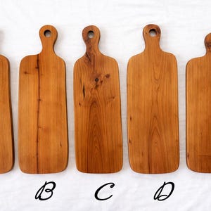 Pu&ograve; includere: Cinque taglieri in legno con finitura naturale venata. Ogni tagliere ha un manico con un foro per appenderlo. I taglieri variano leggermente per forma e dimensione, con una calda tonalit&agrave; marrone-rossastra. I taglieri sono etichettati A, B, C, D ed E.
