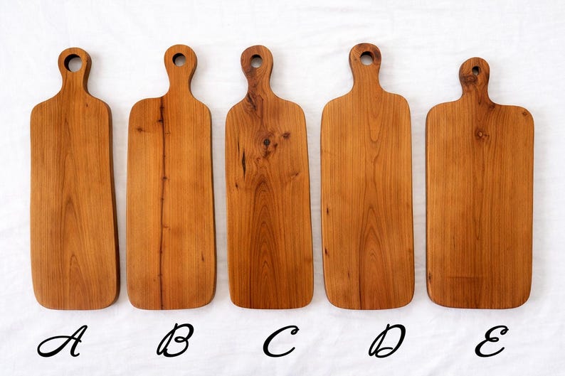 Pu&ograve; includere: Cinque taglieri in legno con finitura naturale venata. Ogni tagliere ha un manico con un foro per appenderlo. I taglieri variano leggermente per forma e dimensione, con una calda tonalit&agrave; marrone-rossastra. I taglieri sono etichettati A, B, C, D ed E.