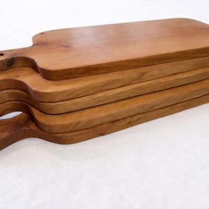 Pu&ograve; includere: Una pila di sei taglieri in legno con manici. Le tavole sono di un caldo colore marrone e hanno una finitura liscia. Ogni tavola ha un manico con un foro per appenderla. Le tavole sono impilate l'una sull'altra.