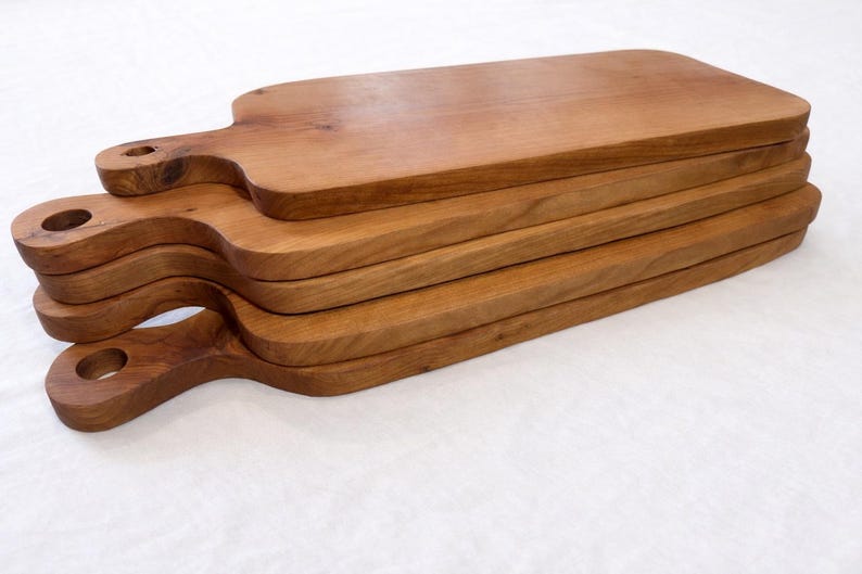 Pu&ograve; includere: Una pila di sei taglieri in legno con manici. Le tavole sono di un caldo colore marrone e hanno una finitura liscia. Ogni tavola ha un manico con un foro per appenderla. Le tavole sono impilate l'una sull'altra.