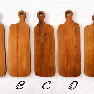 Pu&ograve; includere: Sono presentati cinque taglieri in legno con finitura naturale. Ogni tagliere ha un manico con un foro per appenderlo. I taglieri sono etichettati dalla A alla E. I taglieri variano leggermente nella forma e nella venatura.
