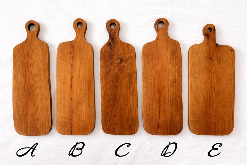 Pu&ograve; includere: Sono presentati cinque taglieri in legno con finitura naturale. Ogni tagliere ha un manico con un foro per appenderlo. I taglieri sono etichettati dalla A alla E. I taglieri variano leggermente nella forma e nella venatura.