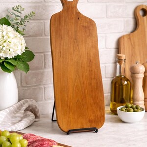 Pu&ograve; includere: Un tagliere in legno con manico, esposto su un supporto in metallo nero. La tavola ha venature naturali del legno e un foro nel manico. Sono visibili anche altri taglieri in legno, una bottiglia di olio d'oliva e una ciotola di olive.