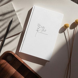 Peut inclure: Un journal blanc avec le texte "MY IVF Planner" et un motif floral. Un crayon noir et un carnet beige sont &agrave; c&ocirc;t&eacute; du journal. Un plateau en bois et des fleurs jaunes s&eacute;ch&eacute;es sont &eacute;galement dans l'image.