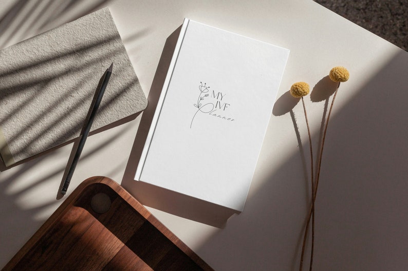 Peut inclure: Un journal blanc avec le texte "MY IVF Planner" et un motif floral. Un crayon noir et un carnet beige sont &agrave; c&ocirc;t&eacute; du journal. Un plateau en bois et des fleurs jaunes s&eacute;ch&eacute;es sont &eacute;galement dans l'image.