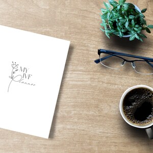Peut inclure: Un cahier blanc avec le texte "MY IVF Planner" et un motif floral, &agrave; c&ocirc;t&eacute; d'une tasse de caf&eacute;, de lunettes, d'une petite plante et d'un ordinateur portable sur une surface en bois. La tasse &agrave; caf&eacute; est noire.