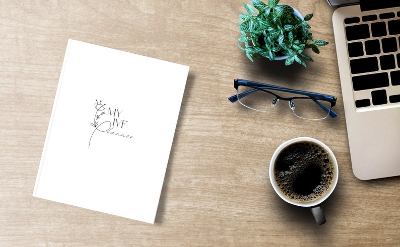 Peut inclure: Un cahier blanc avec le texte "MY IVF Planner" et un motif floral, &agrave; c&ocirc;t&eacute; d'une tasse de caf&eacute;, de lunettes, d'une petite plante et d'un ordinateur portable sur une surface en bois. La tasse &agrave; caf&eacute; est noire.