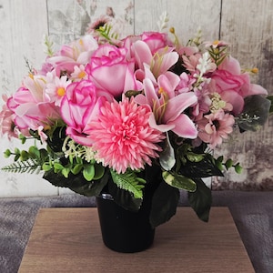 May include: A colourful arrangement of artificial pink flowers, including roses, lilies, and daisies, in a black pot. The flowers have varying shades of pink and green foliage, creating a full display. The pot sits on a wooden surface.