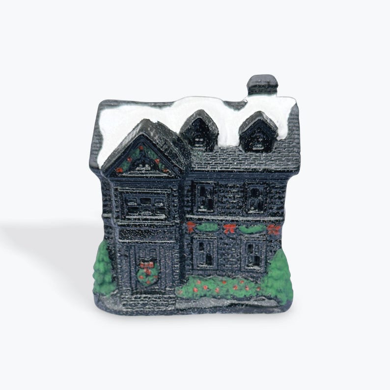May include: A small black and white ceramic Christmas house figurine with a snowy roof and green garland decorations.