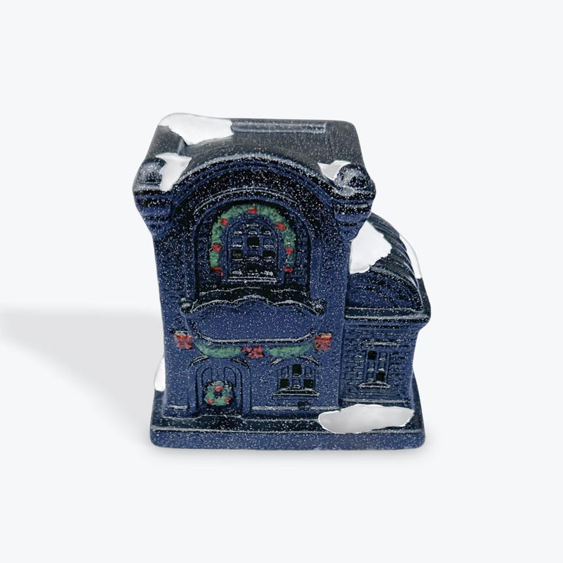 May include: A small, blue ceramic Christmas house with white snow accents. The house has a large window with a wreath and a smaller window with a wreath below it.