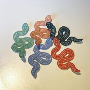 Op de afbeelding: Een verzameling glitterende slangenstickers in verschillende kleuren, waaronder groen, oranje, blauw, zwart en rood. Elke sticker heeft een golvend ontwerp en is bedekt met sprankelende glitter. De stickers zijn gerangschikt op een wit oppervlak.