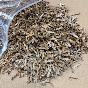 Può includere: Un mucchio di strisce di cartone ondulato marrone sminuzzato che fuoriescono da un sacchetto di plastica trasparente con chiusura a zip. Il materiale sminuzzato è composto da pezzi di carta marrone sottili, stropicciati e piatti con segni di inchiostro.
