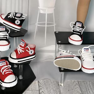 May include: A collection of high-top sneakers in black, white, and red, featuring white laces, white rubber toe caps, and a circular logo on the side with the text Little Step. The shoes are displayed on tiered black acrylic stands.