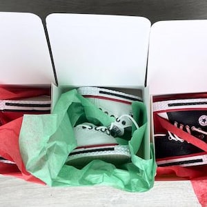 May include: Three white cardboard boxes containing pairs of high-top sneakers. The left and center boxes hold white sneakers with red and black trim, while the right box contains black sneakers with white laces and a circular logo on the side.