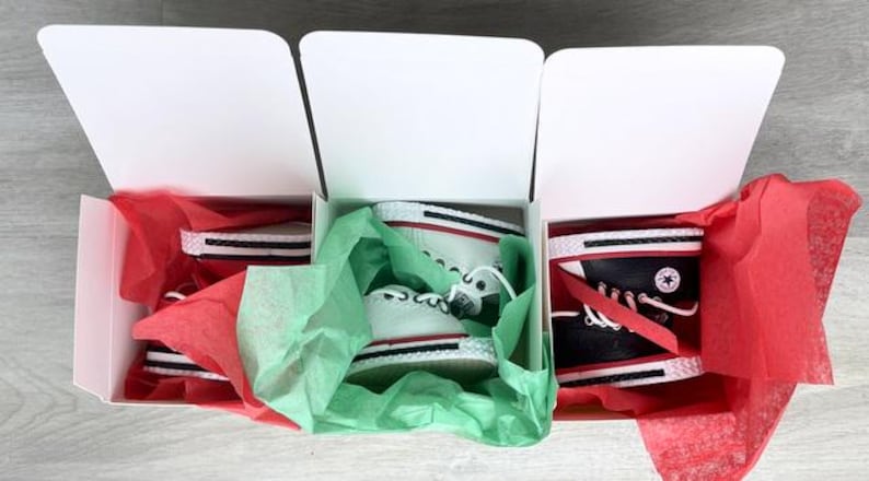 May include: Three white cardboard boxes containing pairs of high-top sneakers. The left and center boxes hold white sneakers with red and black trim, while the right box contains black sneakers with white laces and a circular logo on the side.