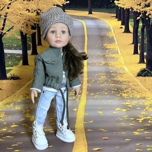 Könnte beinhalten: Eine Puppe mit einer grauen Strickmütze, einer grünen Jacke, einem gestreiften Pullover, hellblauen Jeans und weißen Turnschuhen. Die Puppe hat lange braune Haare und steht auf einer Straße mit gelben Blättern.