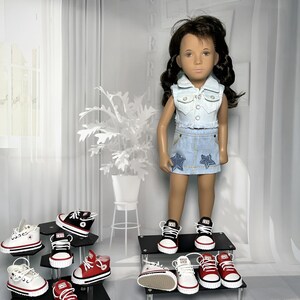 May include: A doll with dark brown hair wearing a light blue denim vest and a matching denim skirt with star appliques. The doll wears black and white sneakers. Several pairs of miniature sneakers in red, white, and black are displayed nearby.