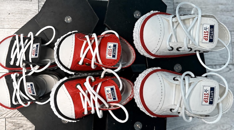 May include: Three pairs of low-top sneakers in black, red, and white. Each shoe features white laces, a white rubber toe cap, and a rectangular tongue label with the text LITTLE STEP and a star icon. The soles have a textured white trim.