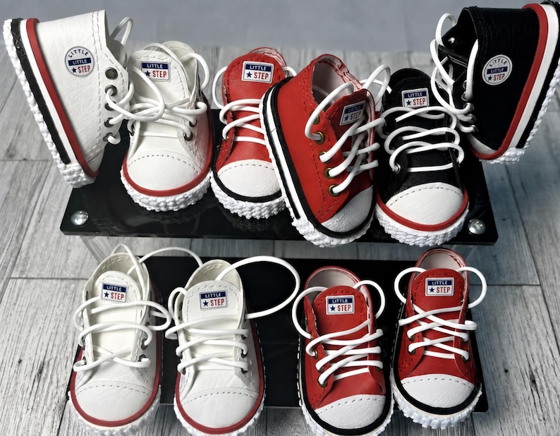 May include: A collection of miniature high-top and low-top sneakers in white, red, and black colors. Each shoe features white laces, a white rubber toe cap, and a circular patch on the side or tongue with the text Little Step and a star icon.