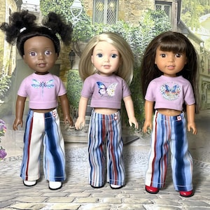 May include: Three dolls standing side by side, each wearing a light purple short-sleeved crop top with a butterfly graphic and long trousers featuring vertical blue, white, and red stripes. The dolls have different hair colours and styles.