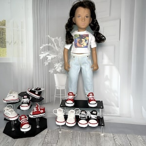 May include: A doll with dark hair in pigtails wears a white t-shirt with a colorful guitar graphic and light blue jeans. The doll stands on a black tiered display stand surrounded by several pairs of miniature canvas sneakers in red, black, and white.