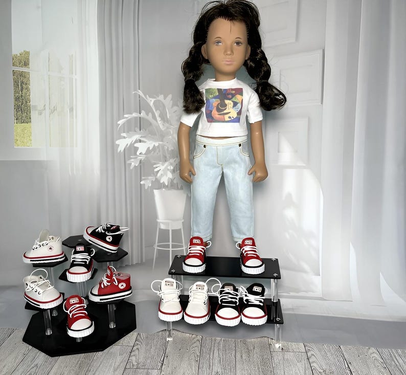 May include: A doll with dark hair in pigtails wears a white t-shirt with a colorful guitar graphic and light blue jeans. The doll stands on a black tiered display stand surrounded by several pairs of miniature canvas sneakers in red, black, and white.
