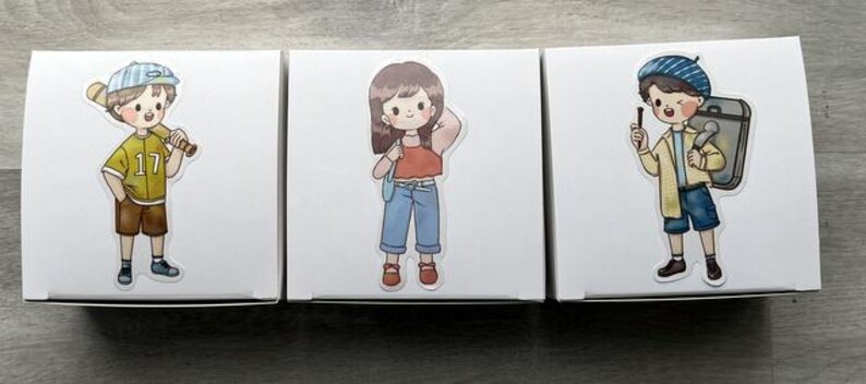 May include: Three white square boxes, each featuring a distinct cartoon sticker. The first shows a person in a yellow jersey with a baseball bat. The second shows a person in a red top and blue jeans. The third shows a person in a blue beret.