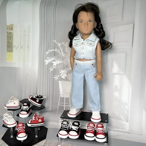 May include: A doll with dark brown hair styled in two braids, wearing a light blue denim vest and matching light blue denim pants. The doll is displayed on a tiered stand alongside several pairs of miniature canvas sneakers in red, black, and white.