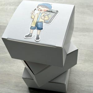 May include: A stack of three plain white square cardboard boxes. The top box features a die-cut sticker of a cartoon character wearing a blue beret, a yellow jacket, and blue shorts, carrying a gray rectangular case and holding a small tool.