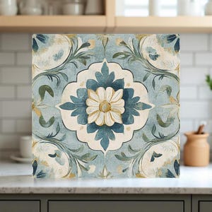 May include: Square decorative tile with a floral design. The tile features a central flower in white and yellow, surrounded by blue and white floral patterns on a light blue background. The tile is likely made of ceramic or porcelain.