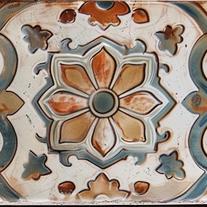 May include: Rectangular ceramic tile with a floral design. The tile features a central flower in shades of orange and tan, surrounded by blue and green scrollwork and accents on a cream background. The tile has a distressed, aged appearance.