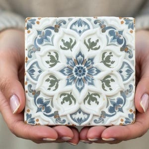 May include: A white ceramic tile with a blue and green floral pattern. The tile has a raised design and is held in the hands of a person.