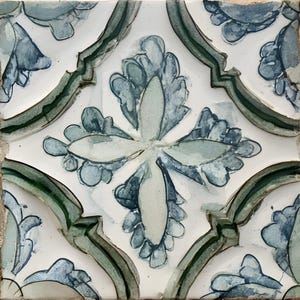 May include: Square ceramic tile with a white background and a repeating floral pattern in shades of blue and green. The design features stylized leaves and a central four-petal flower, with a dark green border.