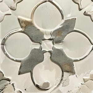 May include: Close-up of a white and gray decorative tile with a repeating floral pattern. The design features four large, circular shapes connected by stylized leaf-like elements. The tile has a glossy finish and a textured appearance.