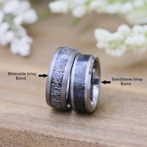 May include: Two tungsten carbide wedding bands with inlay designs. The band on the left has a meteorite inlay, while the band on the right has a sandstone inlay.