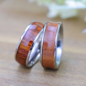May include: Two wooden rings with a silver band around the edge. The rings are made of a dark brown wood with a reddish hue.