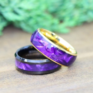 May include: Two men's wedding bands with a purple and gold inlay. The band on top is gold with a purple inlay. The band on the bottom is black with a purple inlay.