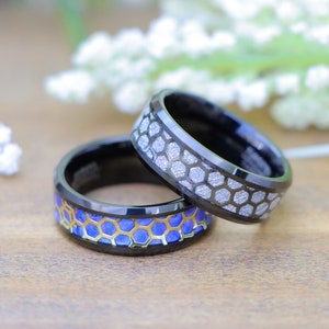 May include: Two black tungsten carbide rings with a honeycomb pattern. One ring has a silver inlay and the other has a blue and gold inlay.