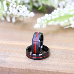 May include: Two black rings with a red stripe. The rings are made of a dark material and have a polished finish. The rings are displayed on a wooden surface with white flowers in the background. The rings are likely wedding bands or fashion accessories.