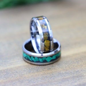 May include: Two silver rings. One ring features a band of green stone chips set in black, while the other has a band of gold-colored stone chips set in black. Both rings have a polished, metallic finish.