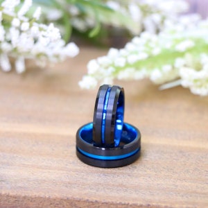 May include: Two black rings with a blue interior and a blue stripe. The rings are on a wooden surface with white flowers in the background. The rings have a modern, sleek design.