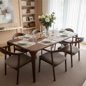 May include: A rectangular wooden dining table with rounded corners and six matching chairs featuring curved backrests and light grey upholstered seats. The table is set with white plates, clear glassware, and a central glass vase of white roses.