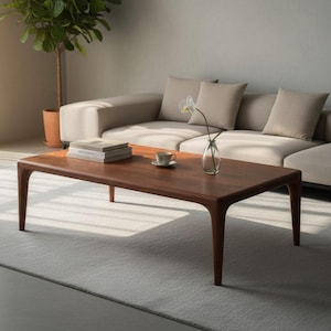 May include: A rectangular wooden coffee table with rounded edges and four tapered legs. The table surface is smooth and dark brown in colour. It is positioned in a room with a light-coloured sofa and a light-coloured rug underneath the table.