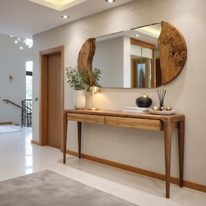 May include: A wooden console table with two drawers, a decorative mirror, and a lamp. The mirror has a unique, organic shape with a wooden frame. A white vase with greenery and a black and gold decorative jar are on the table.