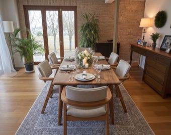 May include: A rectangular wooden dining table with six chairs featuring light-colored upholstered seats and curved wooden frames. The table is set with white plates, silverware, glassware, and a central floral arrangement with three white candles.