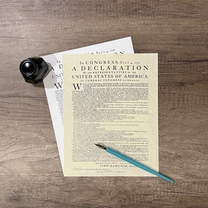May include: A vintage-style print of the Declaration of Independence, framed in a simple white frame. The print is on a wooden surface with a black inkwell and a blue pen.