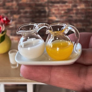 Op de afbeelding: Een wit keramiek dienblad met twee kleine glazen kannen, een gevuld met witte melk en de andere met sinaasappelsap.