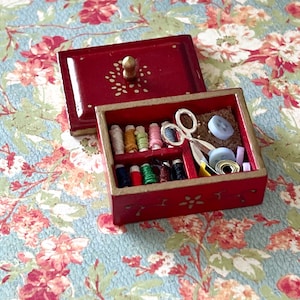 Puede incluir: Una caja de costura de madera roja con ribete dorado y un dise&ntilde;o floral. La caja est&aacute; abierta y contiene carretes de hilo, botones, un par de tijeras y otros art&iacute;culos de costura.