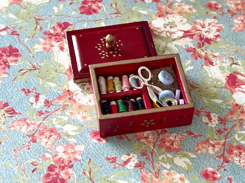 Puede incluir: Una caja de costura de madera roja con ribete dorado y un dise&ntilde;o floral. La caja est&aacute; abierta y contiene carretes de hilo, botones, un par de tijeras y otros art&iacute;culos de costura.